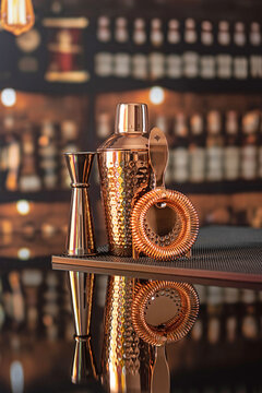 Copper shaker, jigger and strainer on background of bar. Bar accessories are copper-colored. Rubber mat for bar. Set of most popular cocktail mixing equipment. Reflection on table