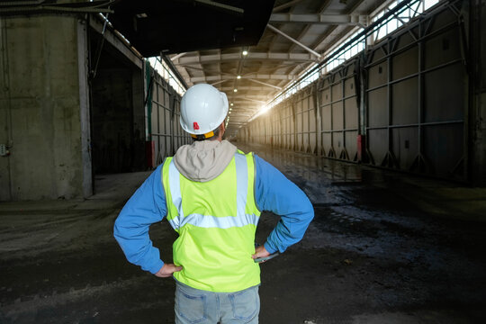 Person in hard hat and safety vest faces away, looking down a long, dark industrial facility hall with sunlight shining in from the far end.