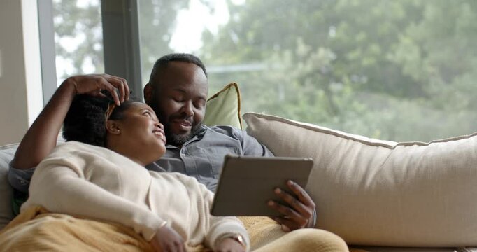African American couple angling tablet, watching while man stroking hair on sofa with yellow throw