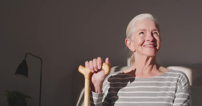 Senior female sitting at home in striped top holding wooden cane, reacting to sunbeam and smiling