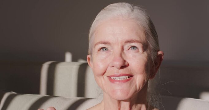 Senior woman squinting from bright sunlight, turning head forward and smiling on sofa in tank top