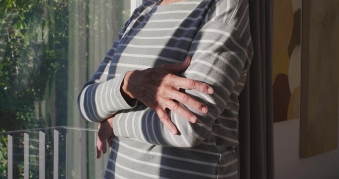 Senior woman feeling sun through blinds adjusting arm lifting chin looking at trees by sliding door