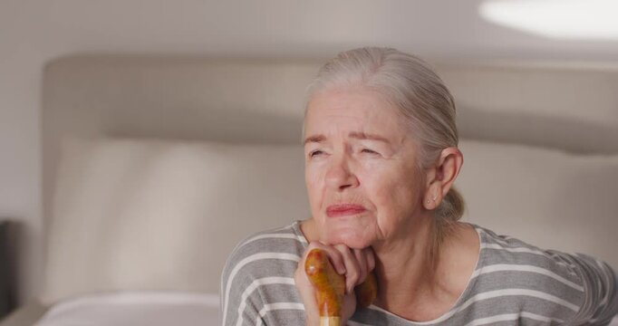 Senior woman on bed resting chin on wooden cane while turning toward bright right light appearing