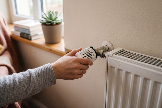 A person is turning down the thermostat on a radiator in a cozy room. The goal is to save energy and reduce heating expenses. A plant is on the window sill nearby