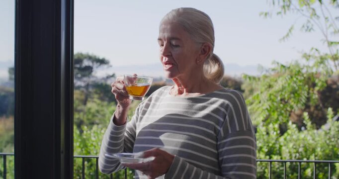Senior woman standing on balcony sipping glass cup and saucer, morning light guiding gaze to hills