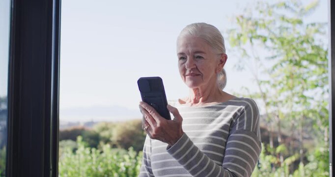 Senior woman standing on balcony wearing striped top with smartphone, scrolling texts and smiling