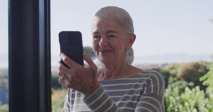 Senior woman raising black phone after text, smiling, laughing at home by glass door, striped top