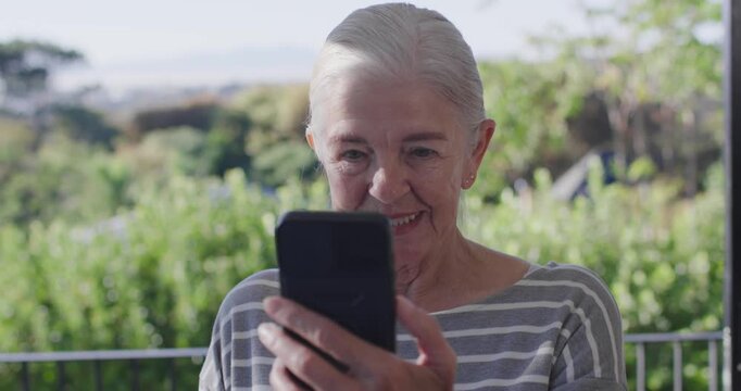 Senior woman raising smartphone, steadying and tapping screen while watching clip on balcony