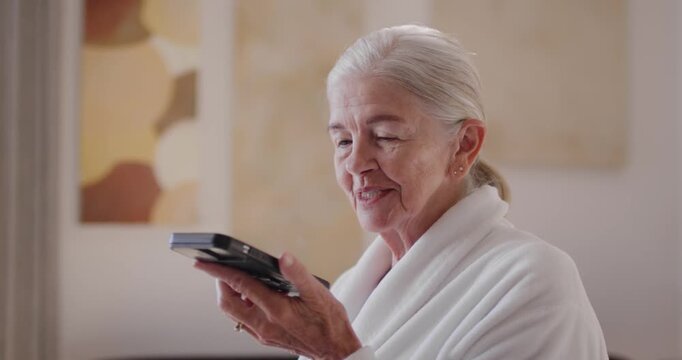 Curious Senior woman lifting handheld device, checking screen, dictating in white bathrobe at home