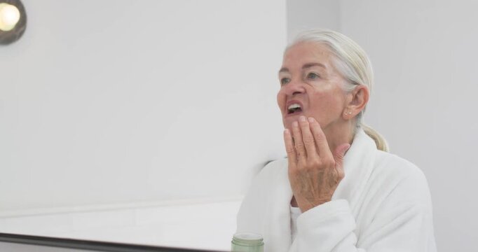 Senior woman in white robe inspecting face at mirror, applying cream, massaging jawline, copy space