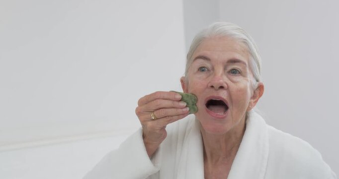 Senior female opening mouth with green gua sha, gliding jaw or under-eye or forehead home for jaw re