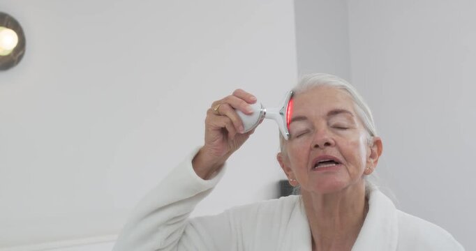 Senior female starting routine in bathroom in bathrobe, holding LED tool glowing red, gliding face