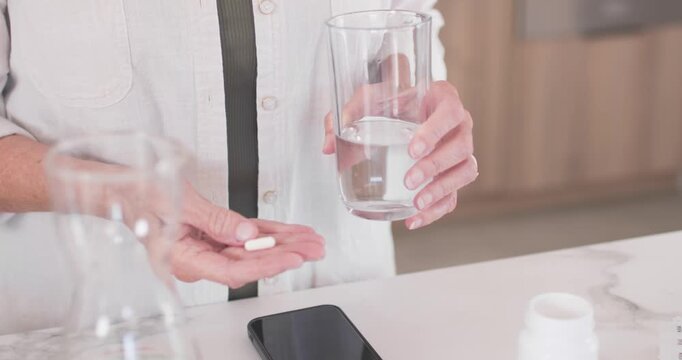 Senior woman holding glass, showing capsules at kitchen counter, shifting pills after seeing bottle