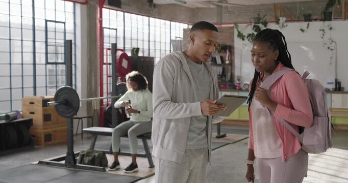 Male staff holding tablet showing plan at gym while female in pink leaning, pointing at tablet