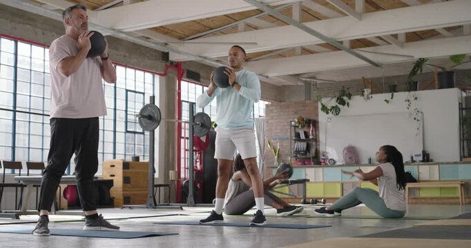 Diverse workout group men squatting with med balls while women alternating core on mats in studio