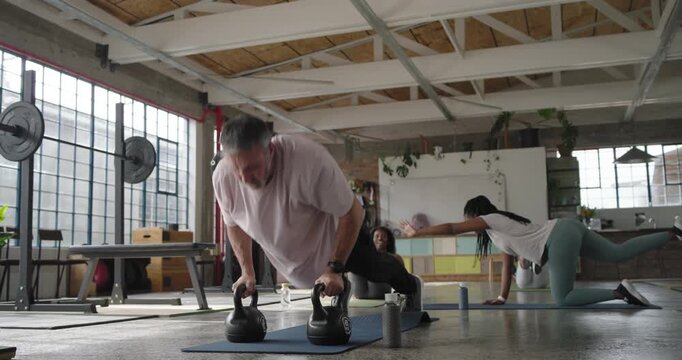 Diverse class after cue doing kettlebell rows on mats by panes, man drinking from bottle recovering