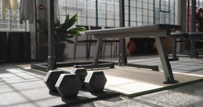 Camera is moving forward and refocusing to highlight three hex dumbbells on rubber mat near bench
