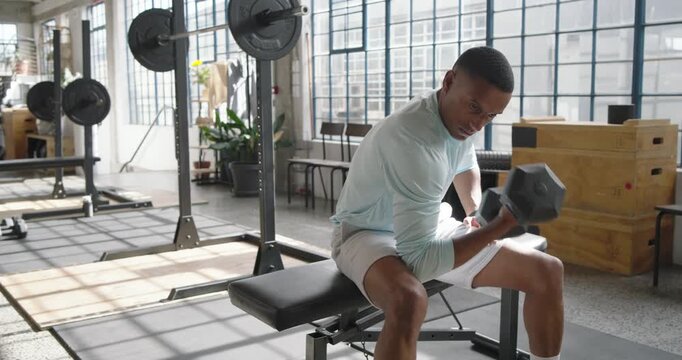 African American man in blue shirt leaning picking hex dumbbell on gym bench, curling for biceps