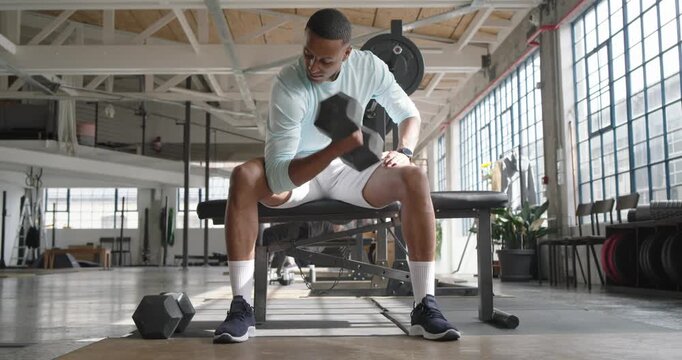 African American man reaching down curling hex dumbbell at gym bench regripping resting toning arms