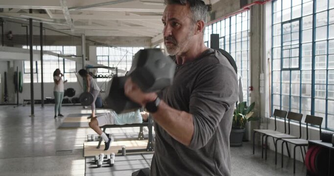 Diverse gym members; mid-40s male doing hex dumbbell curls after pausing, lowering weights, resting