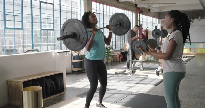 African American coach cueing trainee in turquoise gripping and lifting barbell in gym for strength