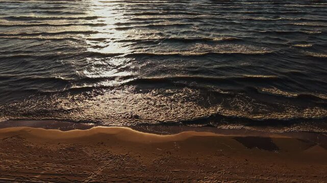 Golden sunset reflection over rippling sea waves beside sandy shoreline and foamy surf