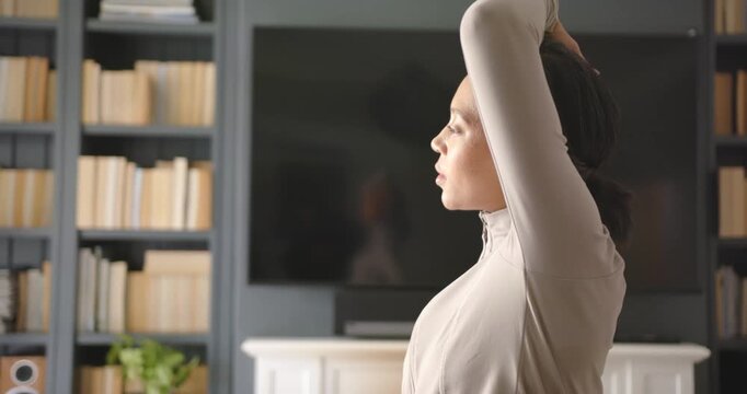 African American woman starting neutral, raising arms behind head holding stretch at home near TV