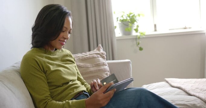 African American woman holding payment card and tapping tablet on couch by window, paying