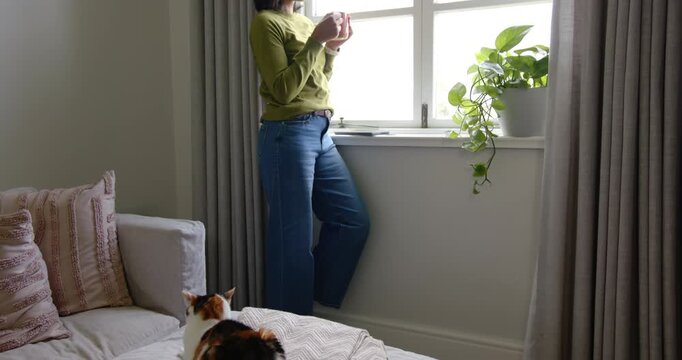 African American woman pulling mug and warming hands, looking at window, cat on bed, copy space