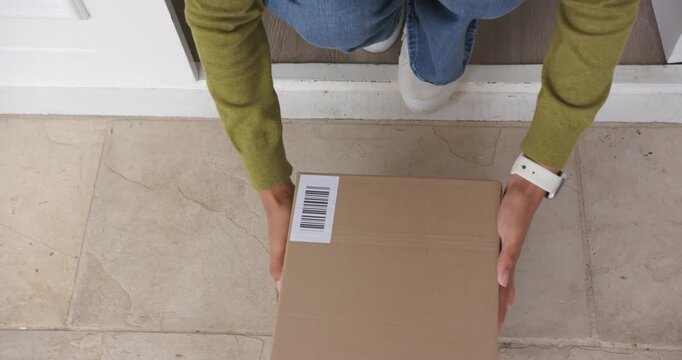 Woman spotting box on threshold, kneeling, grasping barcode-labeled box and carrying it into home