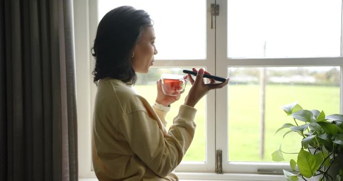African American woman raising smartphone and leaving voice message while sipping tea on windowsill