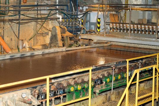 Inside an industrial plant, a large basin holds brown liquid slurry. Numerous pipes, valves, and heavy machinery, including rollers, operate continuously