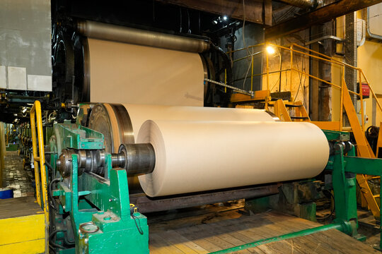 A large sheet of light brown paper moves through industrial machines, winding onto a big roll inside a busy manufacturing plant, showcasing the production process
