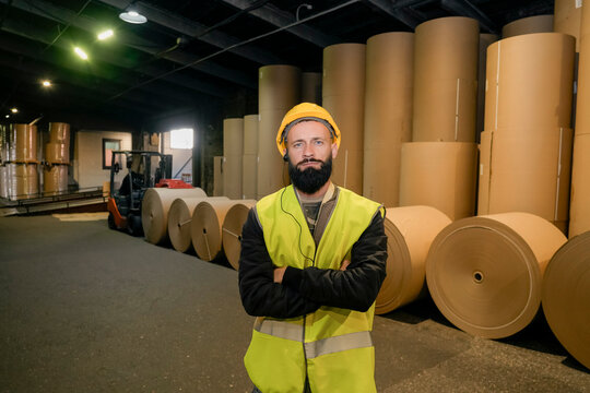 Man with a beard, yellow hard hat, and safety vest stands with crossed arms in a warehouse full of large brown paper rolls and a forklift