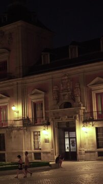 Madrid, Spain, July 4/2026: Night scene of historic building with lit windows and statue. Cobblestone plaza with few people walking. Warm lights highlight architecture and flags