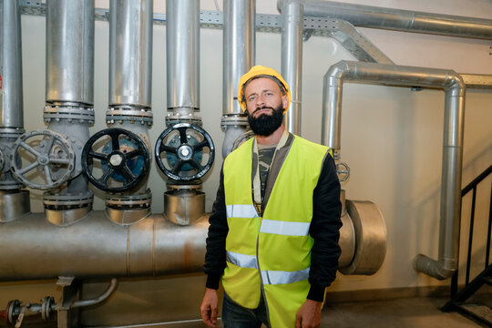 Bearded man in yellow hard hat and bright safety vest stands inside a mechanical room. He is positioned near large silver industrial pipes and control valves