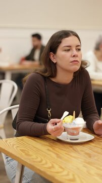 Young woman is sitting at a table in a cafe, eating delicious ice cream and drinking freshly squeezed juice. High quality 4k footage