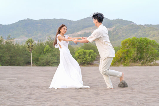 A young Indonesian southeast asian romantic couple in light clothing holding hands and dancing on expansive sand dunes during an outdoor photoshoot in a beautiful landscape