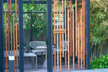 Obraz premium Modern outdoor seating area with comfortable chairs, a glass table, and stylish vertical wooden slats surrounded by green plants for smoking area