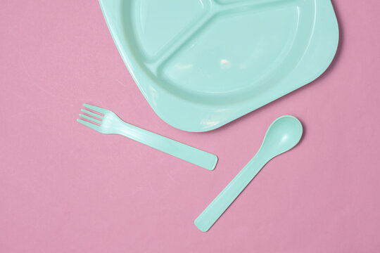 Separate plastic plate with spoon and fork for feeding baby on pink background. Top view