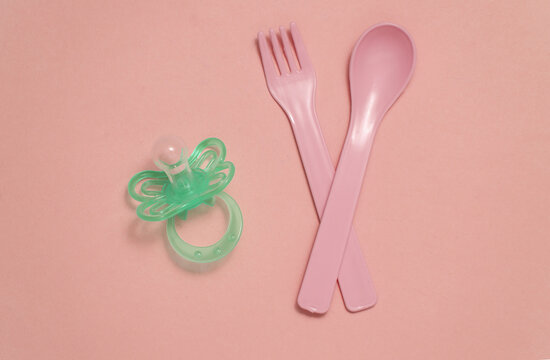 Plastic fork and spoon, pacifier for baby on a pink background. Top view