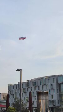 Large airship travels over modern town streets at midday with curious onlookers below