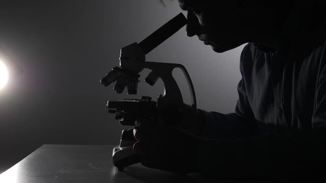 Scientist looking through a microscope, carefully conducting scientific research and analysis in a dark laboratory for discovery and investigation, highlighting innovation in a medical setting