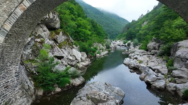 Devil's Bridge in Ardino, Rhodope Mountains, Bulgaria. Low drone flight over the Arda River, passing under the main arch of an ancient stone bridge representative of Ottoman architecture. Europa above