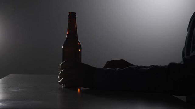 Silhouetted man's hands opening a brown beer bottle against a dark, moody background, conveying themes of loneliness, addiction, and personal struggle with alcohol