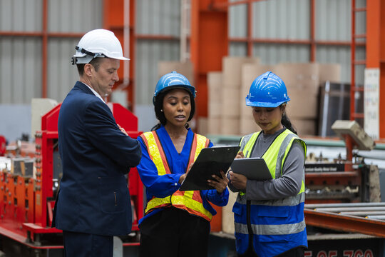 Serious manager and worker team showing diversity during factory inspection discussion wearing safety helmet and vest holding clipboard for industrial production control