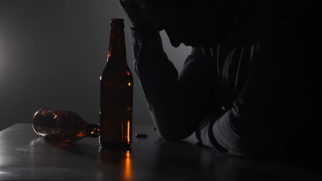 Man covering his face in silhouette, overcome by addiction and depression with empty beer bottles and capsules on a table, symbolizing loneliness and mental health struggles