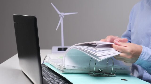 Business person sorting documents for renewable energy planning, using a laptop with a wind turbine model representing green technology and ecological responsibility