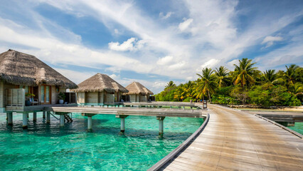 Fototapeta premium Overwater Bungalows Paradise: Serene overwater bungalows dot a crystal-clear turquoise lagoon, connected by a wooden walkway amidst lush tropical vegetation under a bright.