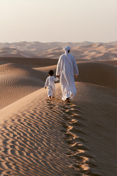 An adult and a child, both in white arabic attire, walking hand in hand over the crest of a sand dune in a sunlit desert.